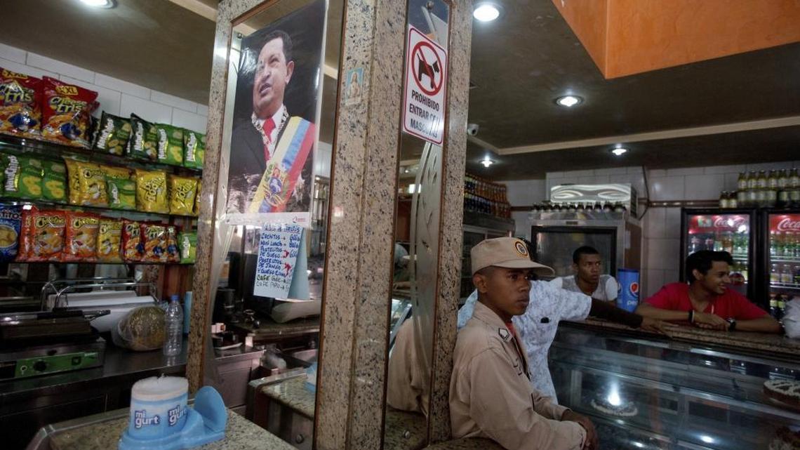 In this March 20, 2017 photo, a militia member stands guard next to a poster of late Venezuelan President Hugo Chavez at a private bakery that was taken over by the government in Caracas, Venezuela. Within hours of the handover, the new storekeepers took down the Coca-Cola-sponsored sign outside and hung up photos of Maduro, Chavez and South American independence hero Simon Bolivar.